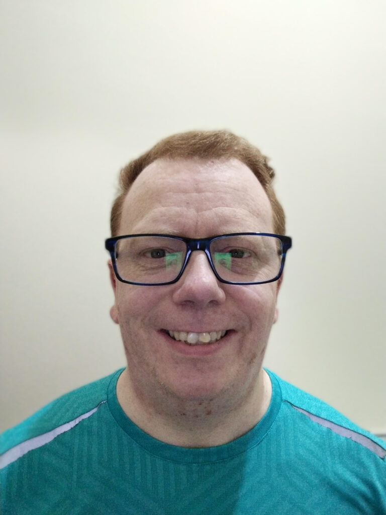 A whit man with short hair, in a blue shirt with blue glasses on.
